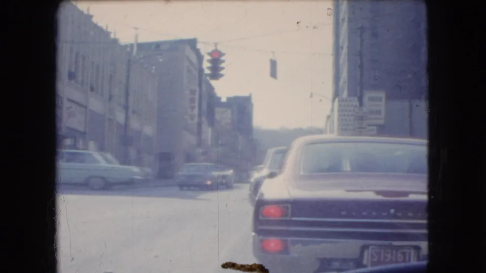 1966 Ohio 8mm frame showing a moving-car view of urban traffic, storefronts, traffic lights, and a brown car ahead in the lane.