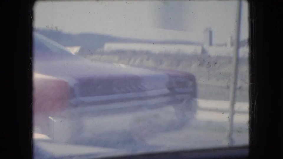 Soft, motion-blurred 8mm frame of a red mid-1960s car passing close to the camera, with roadway and roadside forms in the background.