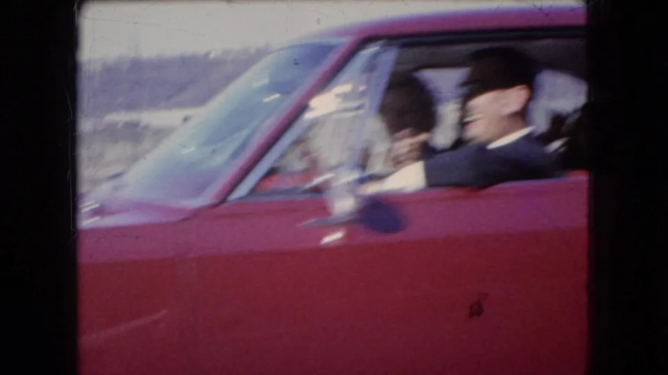 Close 8mm view of a red car in Ohio in 1966, with an adult male driver smiling and another passenger visible beside him.