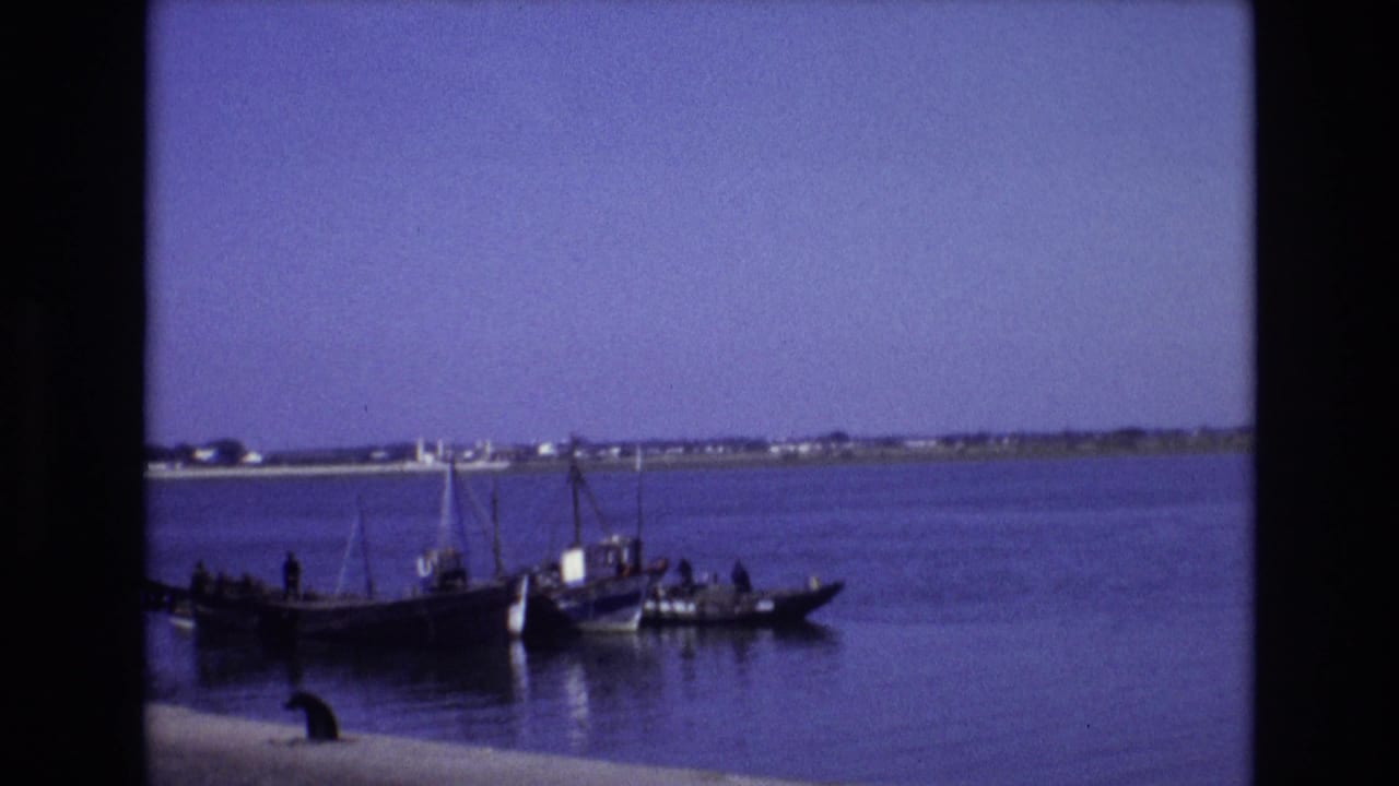 1972 Portugal 8mm frame showing a small dark boat with people aboard on calm blue water, with a distant low shoreline and clear sky behind it.