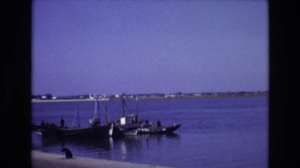 1972 Portugal 8mm frame showing a small dark boat with people aboard on calm blue water, with a distant low shoreline and clear sky behind it.