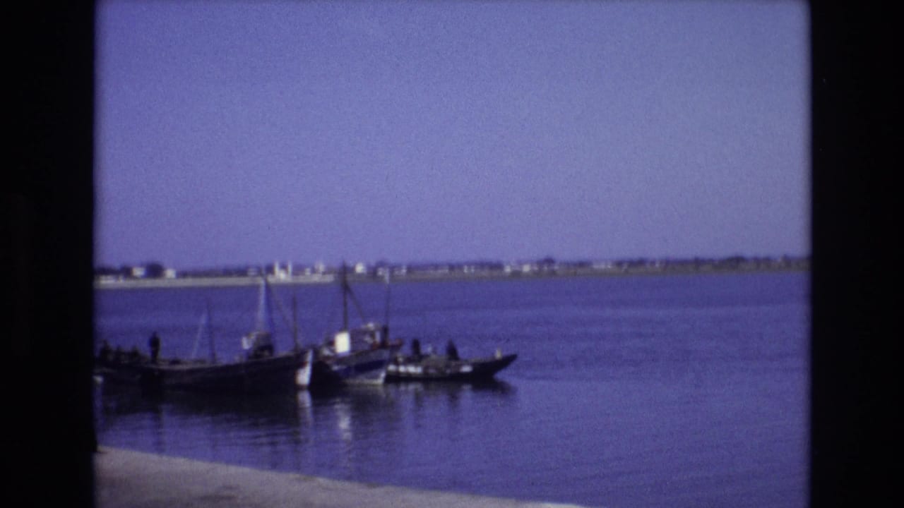 1972 Portugal 8mm frame of a small boat carrying several people on still blue water, with low buildings visible along the far shoreline.