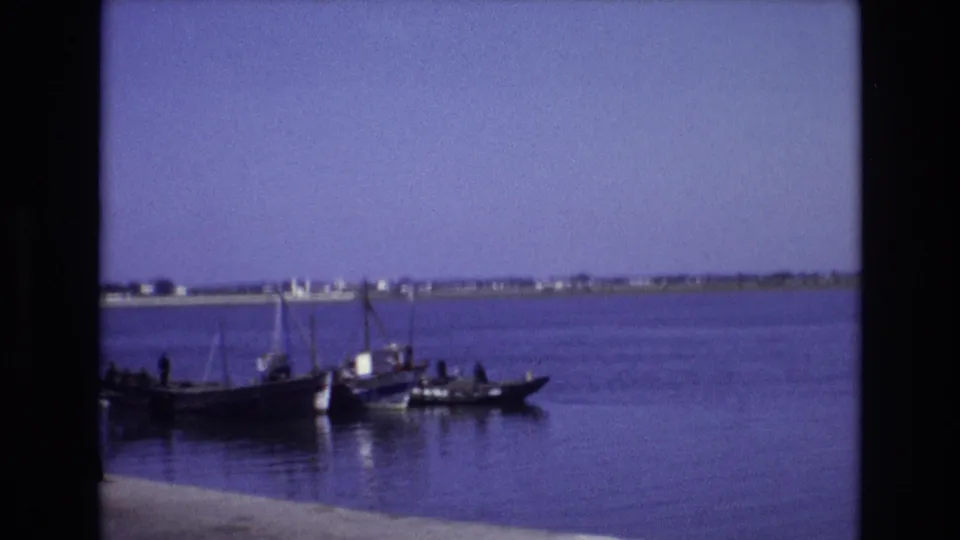 1972 Portugal 8mm frame of a small boat carrying several people on still blue water, with low buildings visible along the far shoreline.