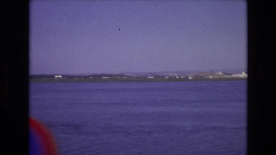 1972 Portugal home-movie frame showing a wide sweep of coastal water, distant shore, and a small reddish edge artifact or foreground intrusion.