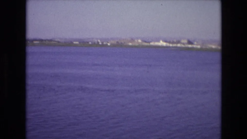1972 Portugal coastal home-movie frame with calm blue water, faint ripples, and a distant shoreline across the horizon.