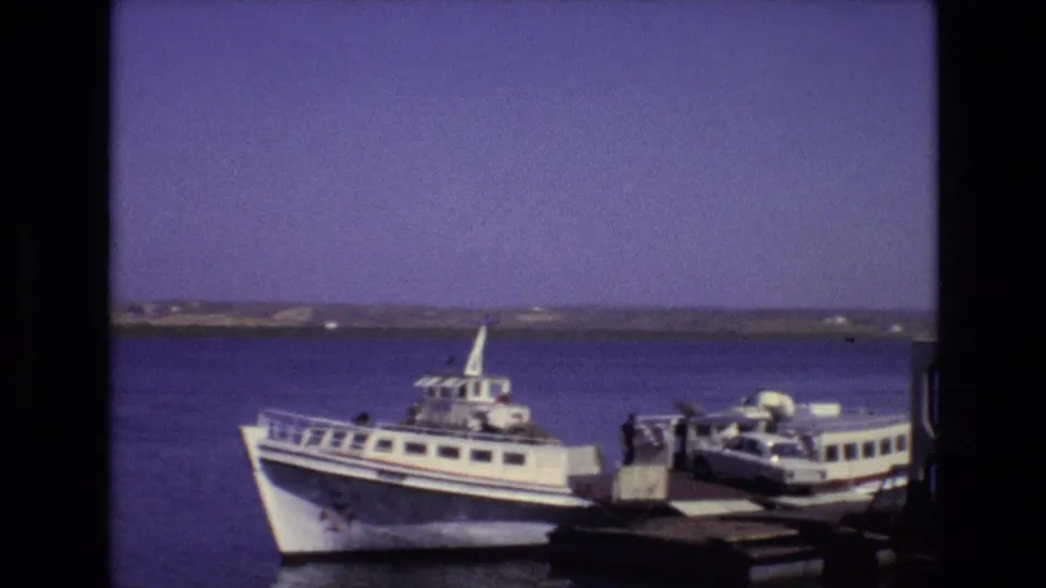 Portugal 1972 ferry scene with a white passenger vessel at a dock, a small car near the loading platform, and calm water behind it.