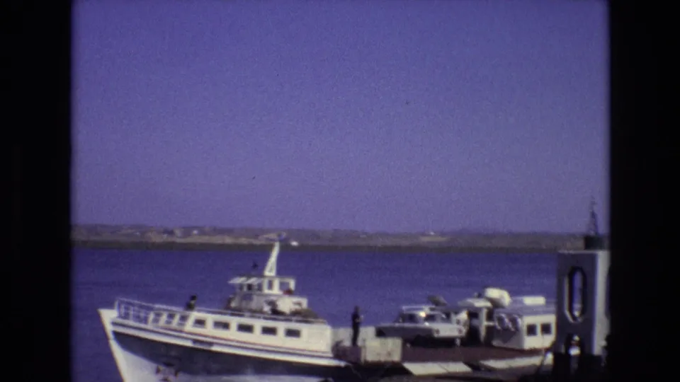 Portugal 1972 home-movie frame with a ferry at the dock, a car beside the ramp, small figures on the platform, and a low shoreline across the water.