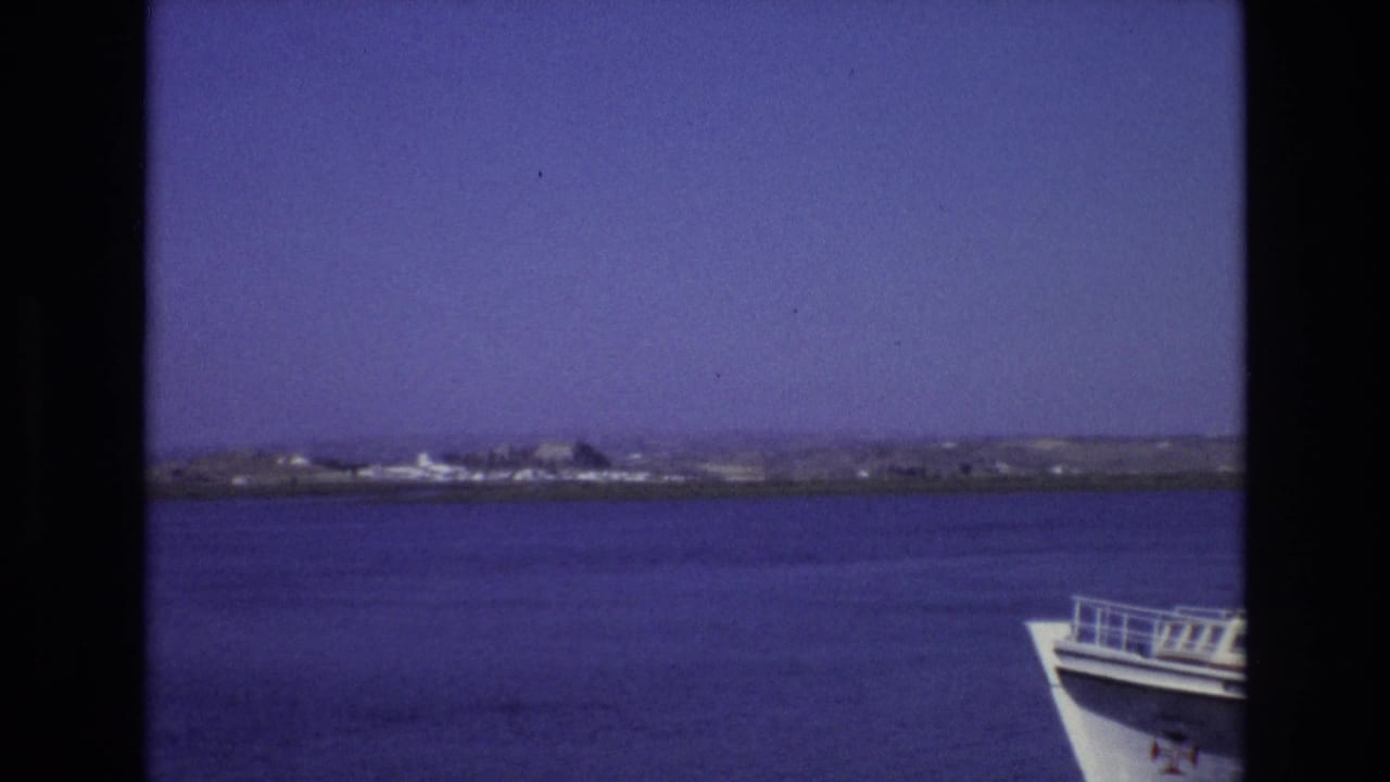 A 1972 Portugal boat-trip view shows open water, a low shoreline, clear sky, and the white bow of the vessel at the right edge.