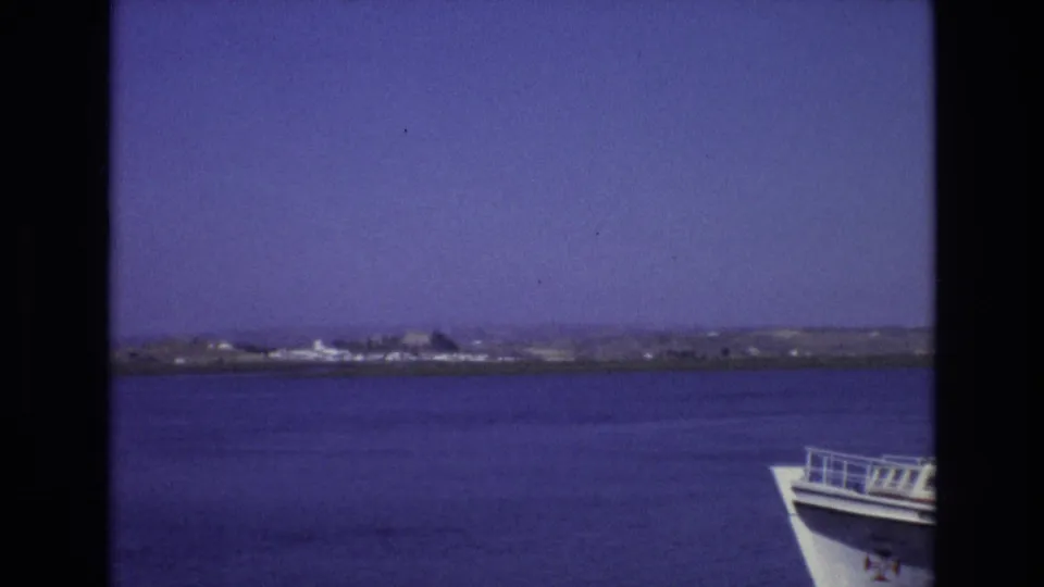 A 1972 Portugal boat-trip view shows open water, a low shoreline, clear sky, and the white bow of the vessel at the right edge.
