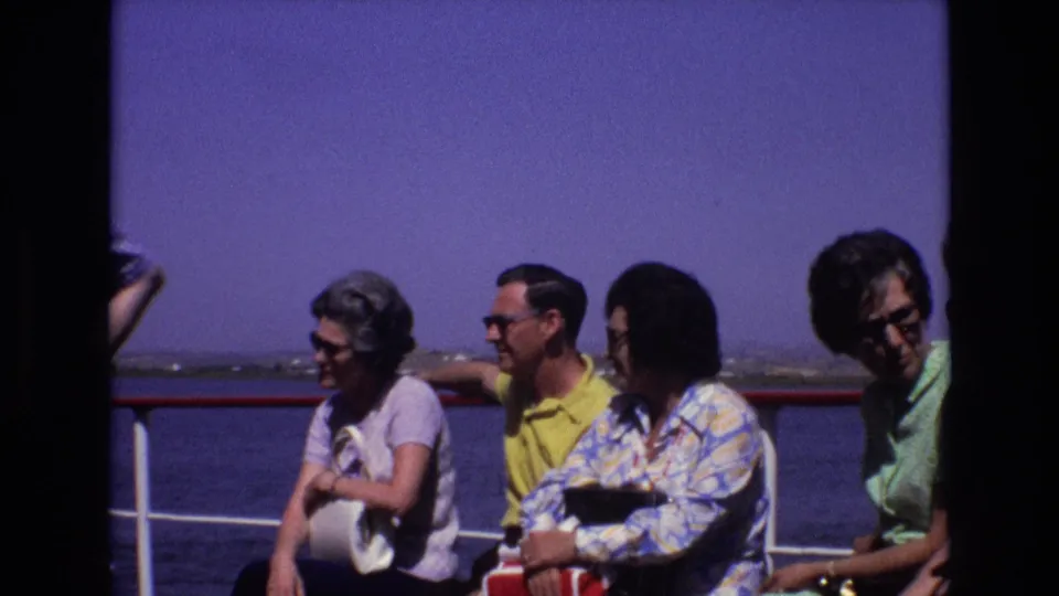 Four adults in 1970s casual clothing sit on a boat deck in Portugal, with water, railing, and bright sky behind them.