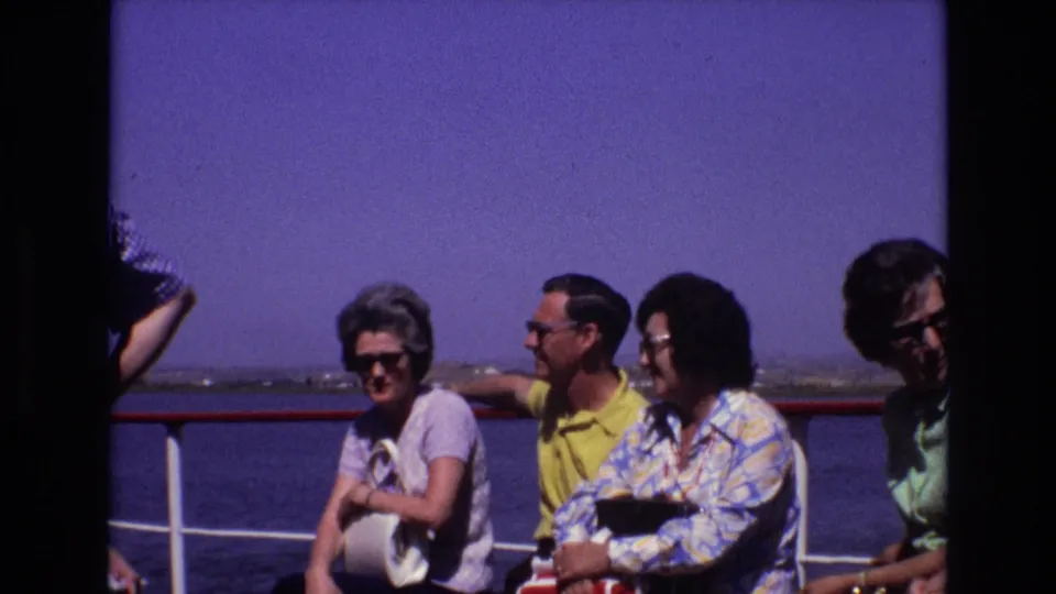 A group of adult tourists sit closely together on a boat deck during a 1972 Portugal outing, with railings, water, and open sky visible behind them.