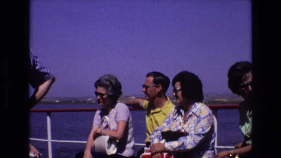 1972 Portugal home-movie frame showing adult passengers in sunglasses seated near a red-and-white ship railing with open water behind them.