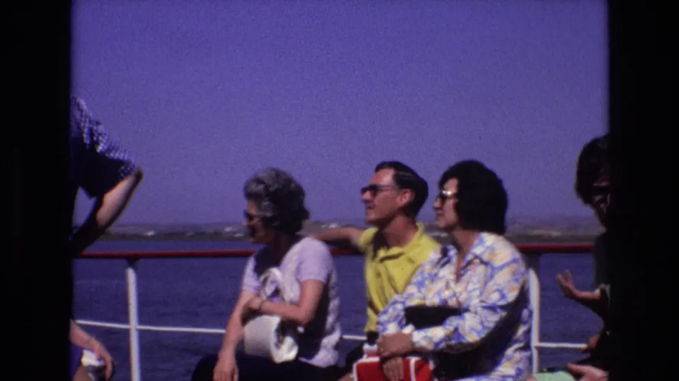 Home-movie view of adult ship passengers seated by the railing, with one woman looking down and the ocean stretching behind the deck.