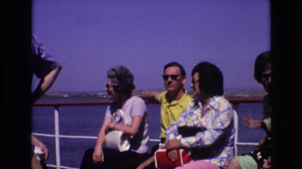 Adult passengers on a ship deck in 1972 Portugal sit near the red-and-white railing while a low shoreline is visible beyond the water.