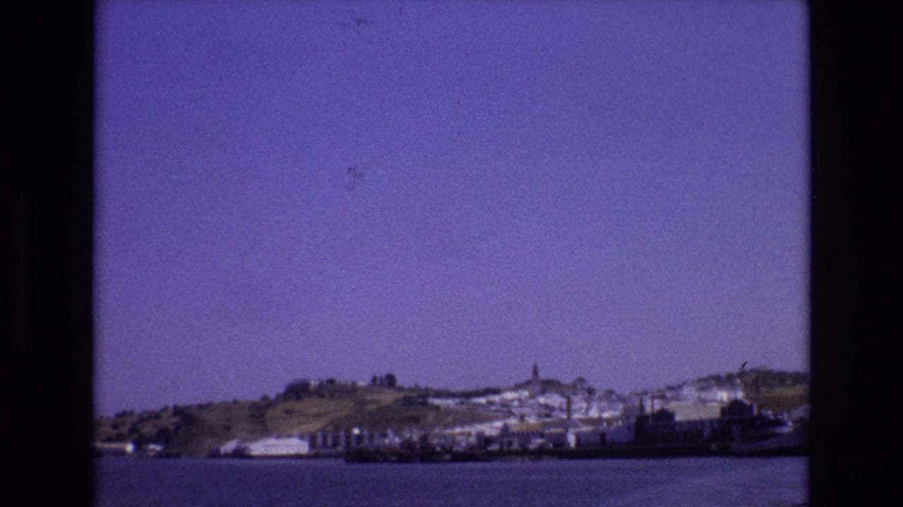 1972 Portugal 8mm frame showing a distant hillside town, shoreline buildings, boats, and calm water under a purple-tinged sky.