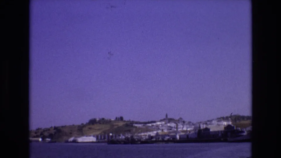 1972 Portugal 8mm frame showing a distant hillside town, shoreline buildings, boats, and calm water under a purple-tinged sky.