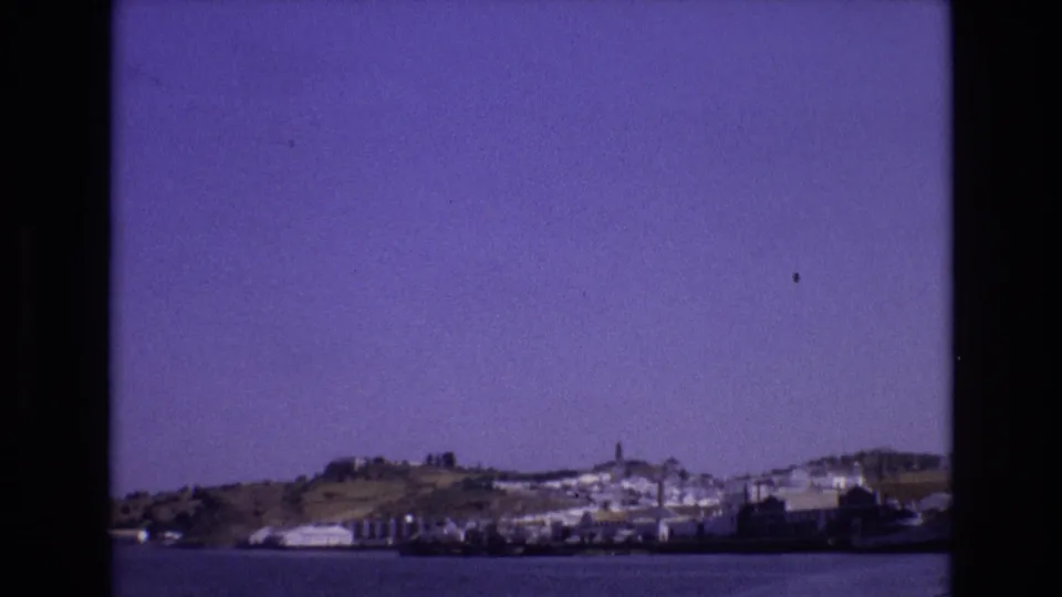 1972 Portugal home-movie still with a broad water foreground, a hillside coastal settlement, moored vessels, and a tower on the skyline.