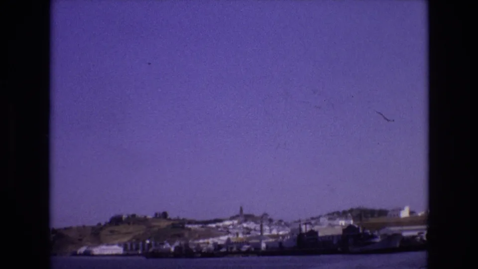 1972 Portugal 8mm view across calm water toward a low waterfront, hillside buildings, a tower, and dark film-frame edges.