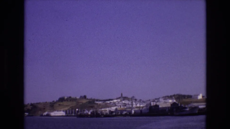 1972 Portugal home-movie frame showing a coastal town, hillside, tower, shoreline vessels, and rippled water from a boat-view perspective.