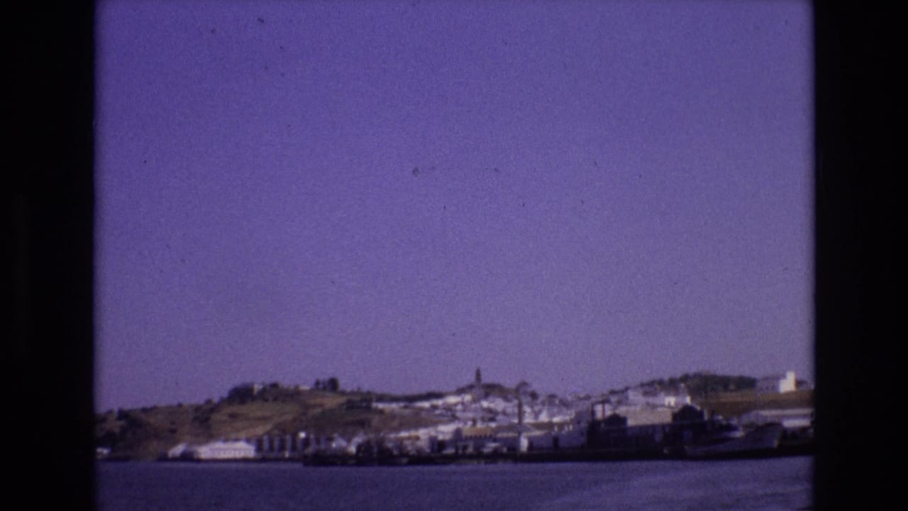 1972 Portugal 8mm frame showing a distant coastal town across the water, with white buildings, a hillside, and a tower visible through dark film borders.