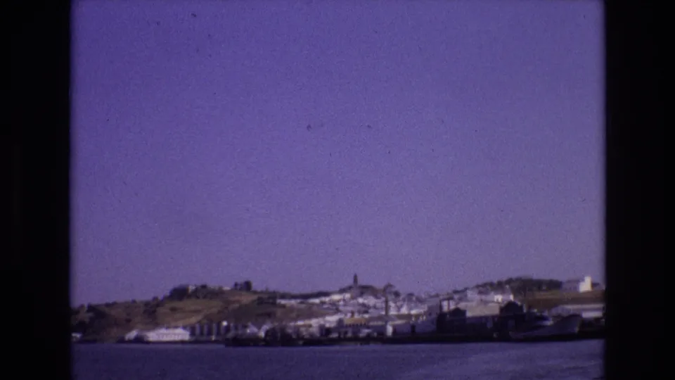 1972 Portugal 8mm frame showing a distant coastal town across the water, with white buildings, a hillside, and a tower visible through dark film borders.