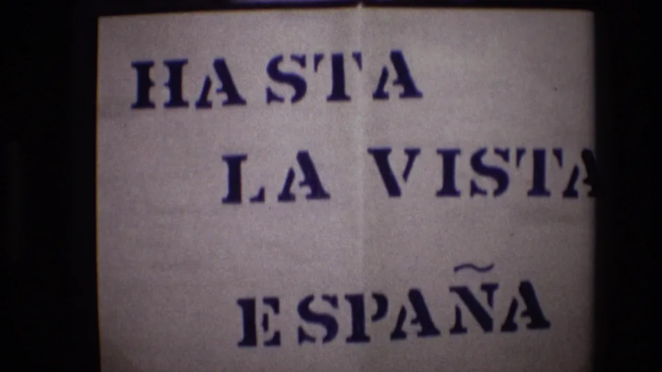 Portugal 1972 8mm frame showing an off-white handmade sign with blue stenciled text reading “HASTA LA VISTA ESPAÑA,” partly framed by dark film borders.