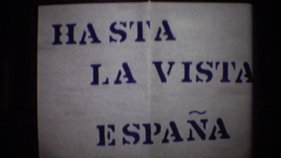 Portugal 1972 8mm frame holding on the handmade sign, with “HASTA LA VISTA” above “ESPAÑA” in bold blue stenciled lettering.