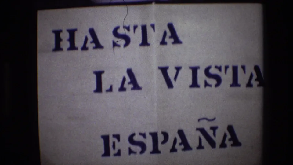 Final 1972 Portugal frame of the creased white paper sign with bold blue HASTA LA VISTA ESPAÑA lettering, soft 8mm grain, and black frame margins.