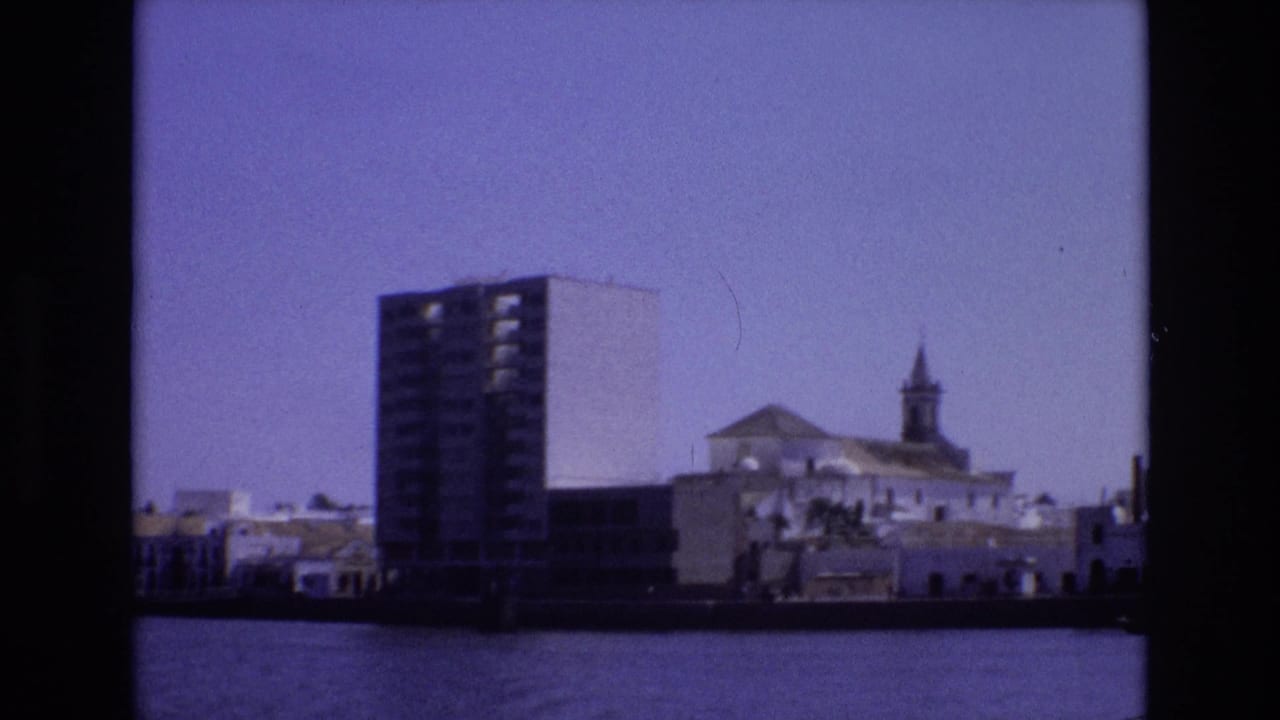 1972 Portugal home-movie frame showing a waterfront cityscape with a tall modern building, low white structures, and a church steeple across calm water.