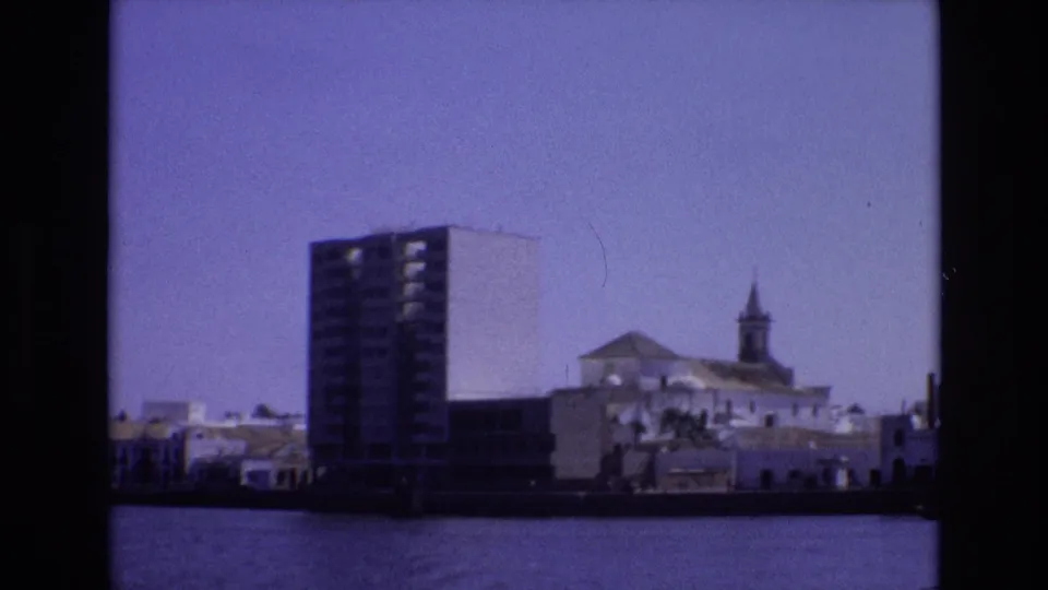 1972 Portugal home-movie frame showing a waterfront cityscape with a tall modern building, low white structures, and a church steeple across calm water.