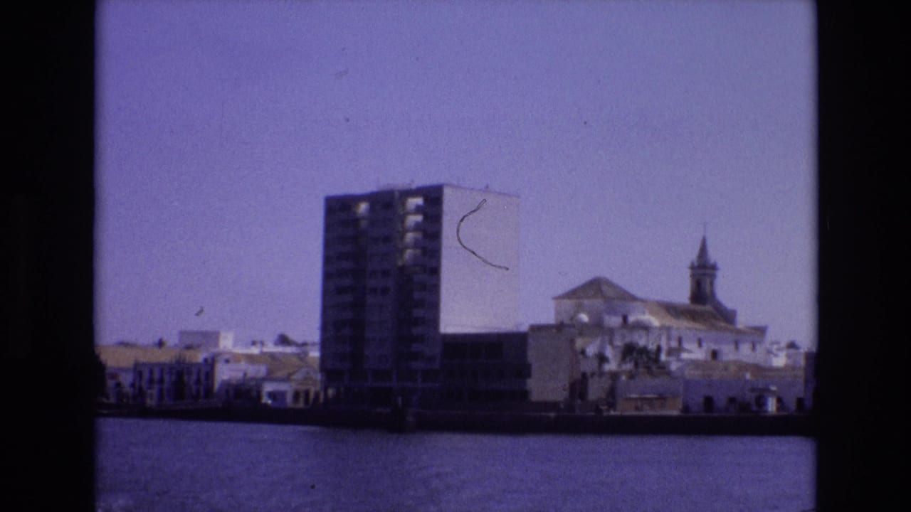 1972 Portugal 8mm frame with a modern high-rise on the waterfront beside white buildings and a church-like structure under a hazy sky.