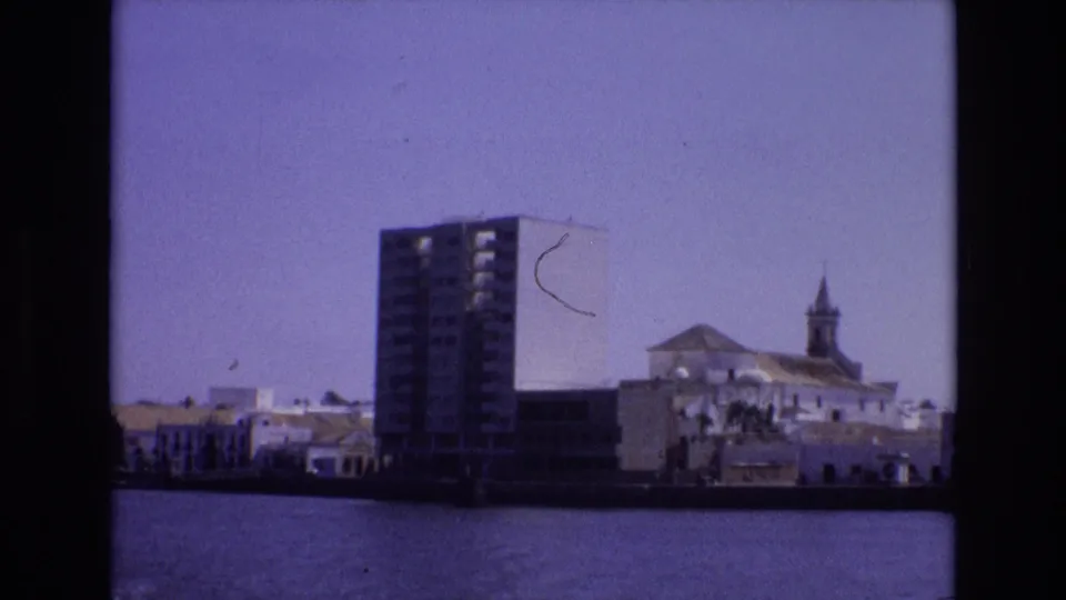 1972 Portugal 8mm frame with a modern high-rise on the waterfront beside white buildings and a church-like structure under a hazy sky.