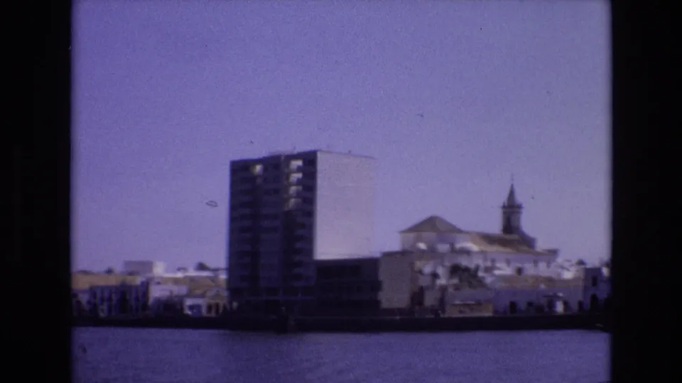 Portugal 1972 home-movie view across water toward a tall block, low white waterfront buildings, and a church steeple softened by 8mm grain.