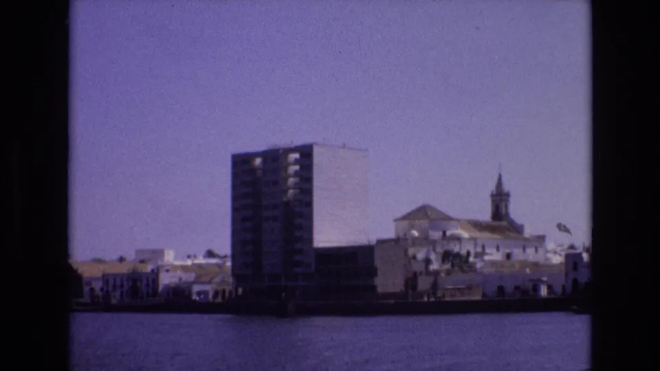 1972 Portugal 8mm frame looking across a waterway toward a modern high-rise beside white waterfront buildings and a church steeple.