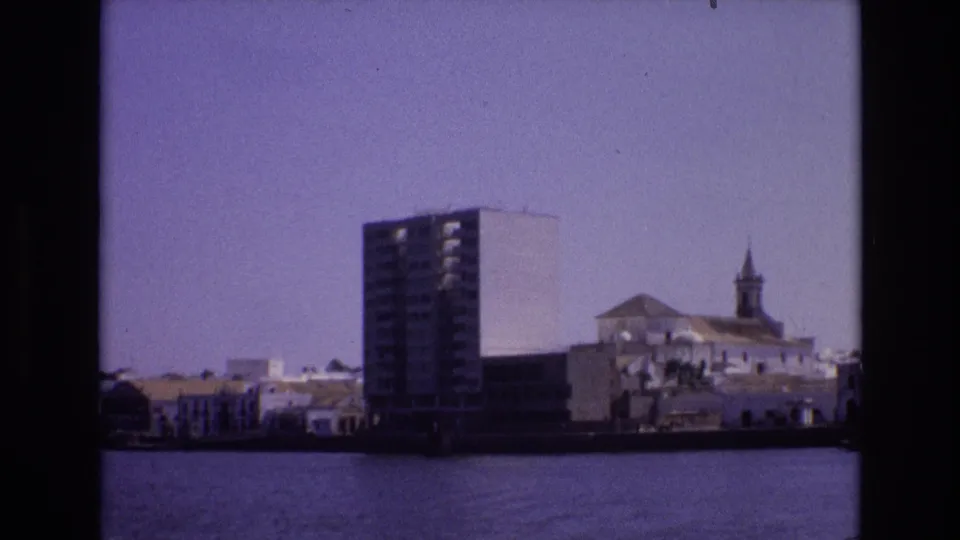 1972 Portugal waterfront view from across the water, with a tall apartment block, white low buildings, and a church steeple visible through soft 8mm grain.