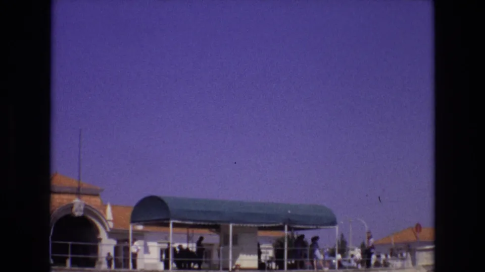 1972 Portugal 8mm frame showing a green-roofed ferry or port canopy with railings, tiled roofs, and distant visitors waiting near the waterfront.