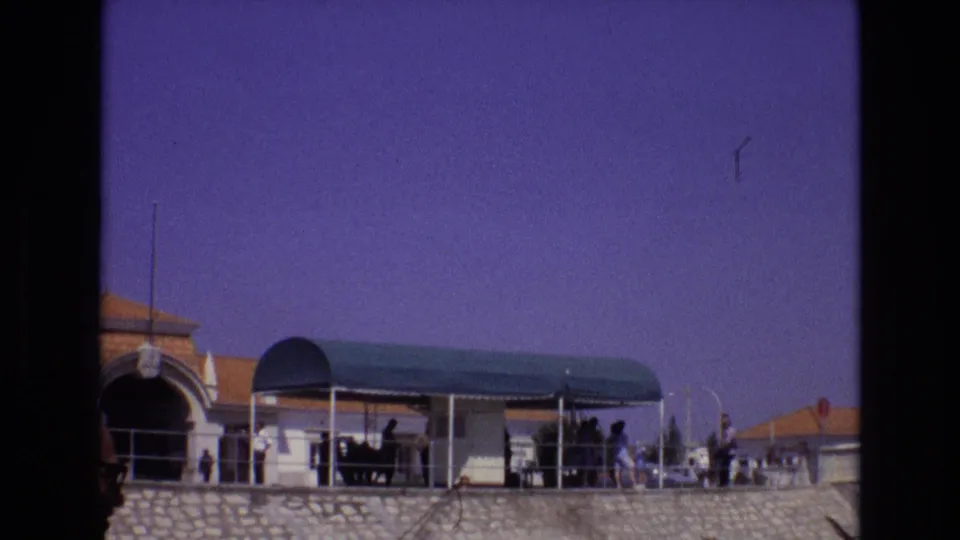 1972 Portugal waterfront landing with visitors gathered near a green canopy, railings, tiled roofs, and a stone embankment in a purple-tinted 8mm frame.