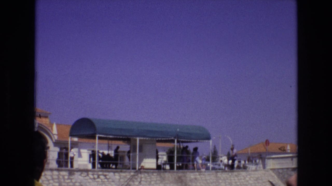 People standing and walking under a green canopy on a stone terrace beneath a blue-purple sky.