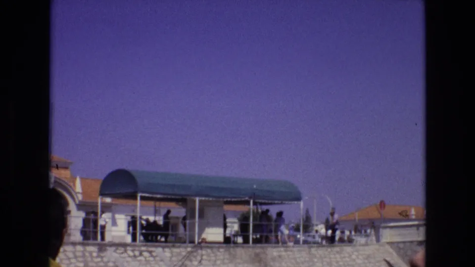 1972 Portugal home-movie frame showing a green arched canopy on a stone terrace, with distant people gathered in shade and terracotta roofs behind them.