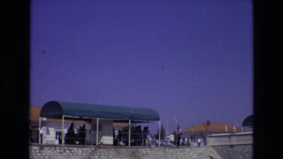 1972 Portugal home-movie frame showing the green terrace canopy lower in the composition, with people in shade, stonework below, and a red roadside sign near terracotta roofs.