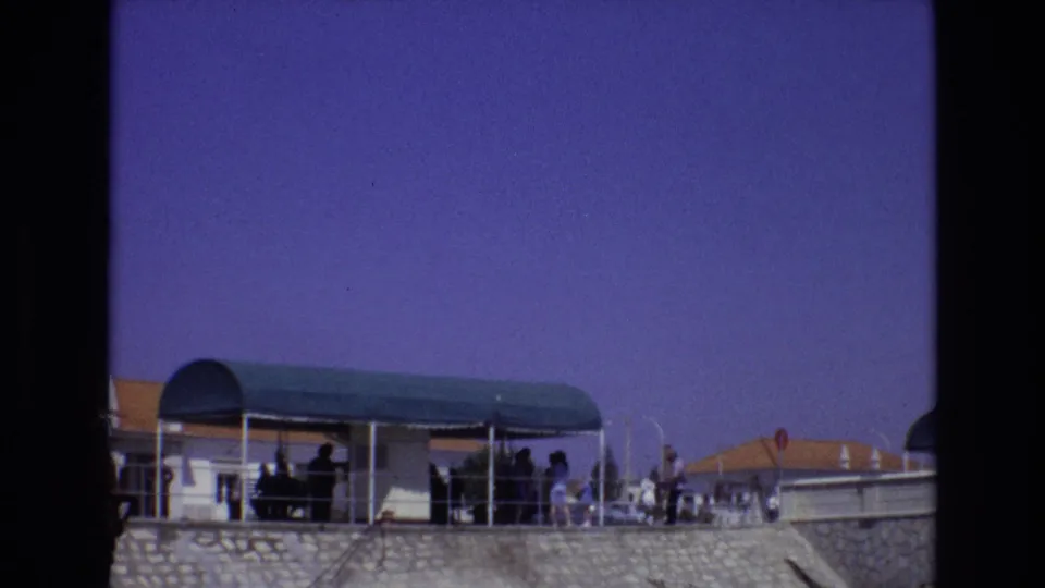 1972 Portugal frame showing distant visitors on a sunlit terrace, a green arched canopy, metal railings, terracotta rooflines, and blue-purple 8mm color shift.