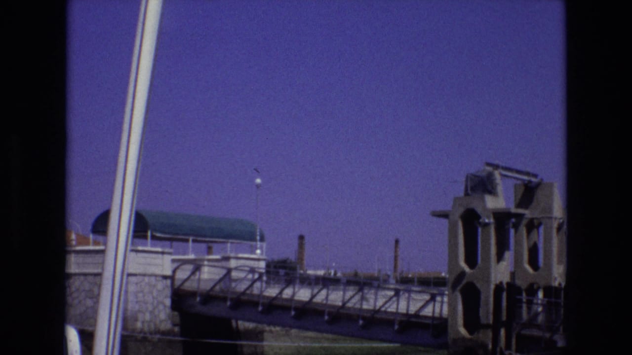 1972 Portugal 8mm frame showing a tilted bridge or walkway view with a concrete tower, stone retaining wall, vertical pale poles, and purple-blue sky.
