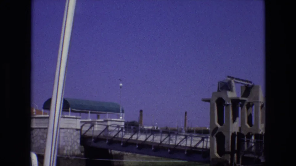 1972 Portugal 8mm frame showing a tilted bridge or walkway view with a concrete tower, stone retaining wall, vertical pale poles, and purple-blue sky.