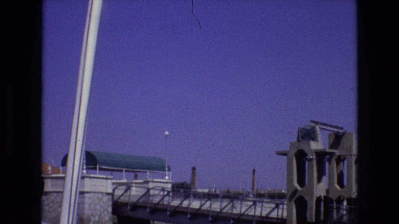 Bridge walkway with small silhouetted people, concrete tower, stone wall, and open sky.