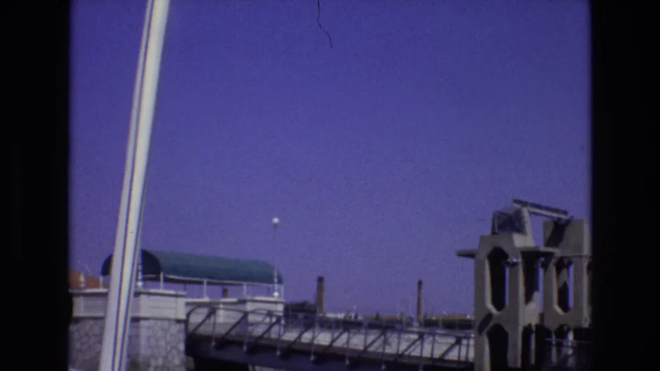 1972 Portugal 8mm frame with a bridge walkway, small silhouetted figures along the railing, a concrete tower with arched openings, stonework, and clear purple-blue sky.