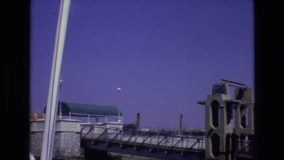 1972 Portugal 8mm frame showing a slightly lower tilted composition of the bridge walkway, concrete tower, stone wall, pale vertical poles, and violet-blue sky.