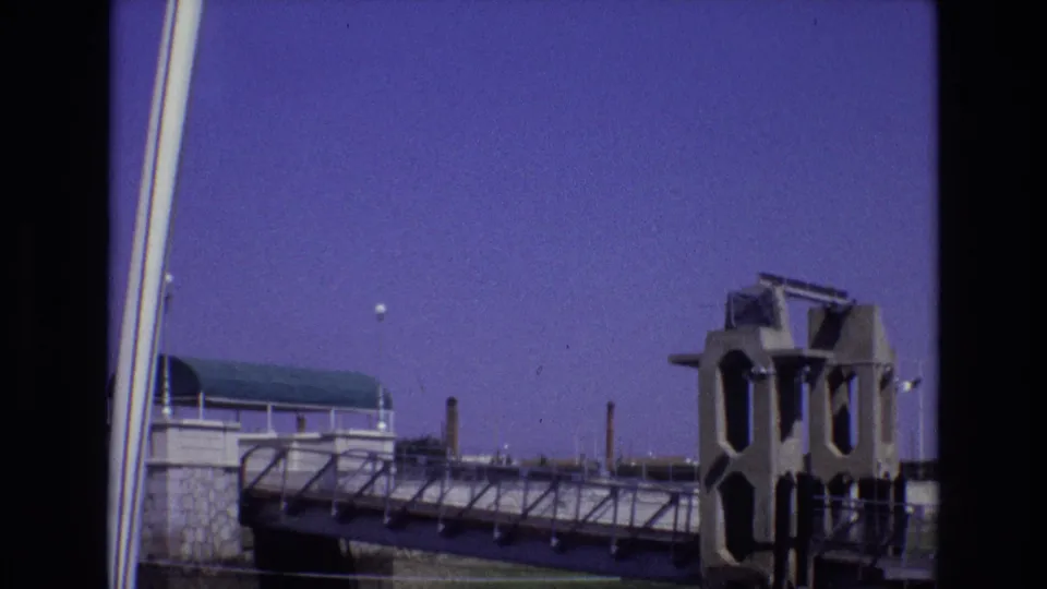 1972 Portugal 8mm frame showing the bridge walkway from a tilted angle, with small silhouetted people near the railing, a concrete tower, stone wall, vertical poles, and purple-blue sky.