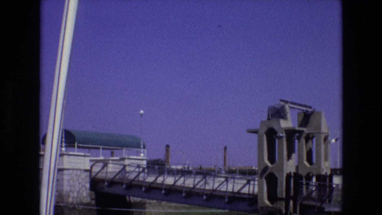 1972 Portugal 8mm frame showing a waterfront or nautical structure with railings, a green-roofed pavilion, industrial stacks, and visible film-frame borders.