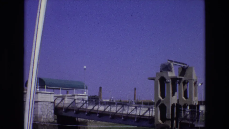 1972 Portugal 8mm frame showing a waterfront or nautical structure with railings, a green-roofed pavilion, industrial stacks, and visible film-frame borders.