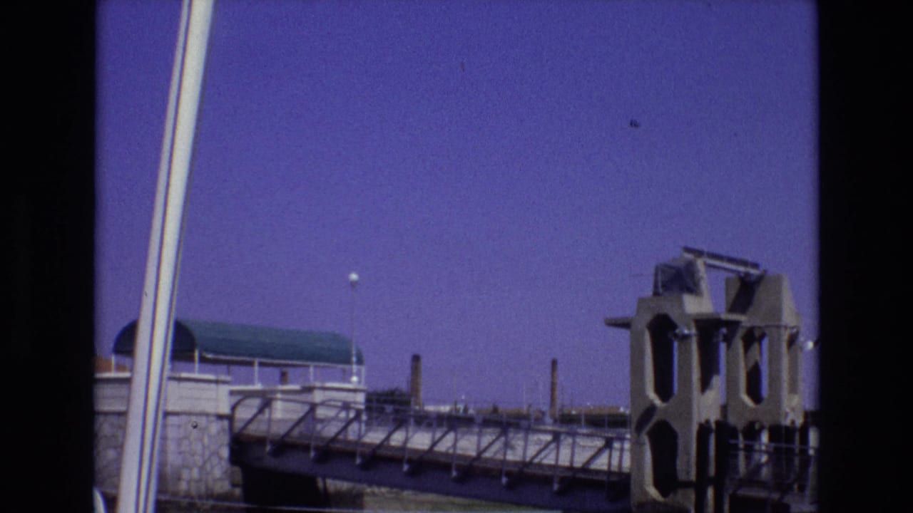 Similar waterfront view with railings, pavilion roof, smokestacks, and a large structural element at right.